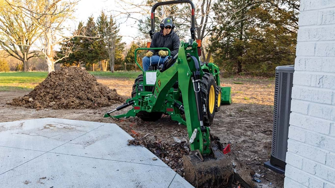 A green backhoe being used to dig a hole near a patio.