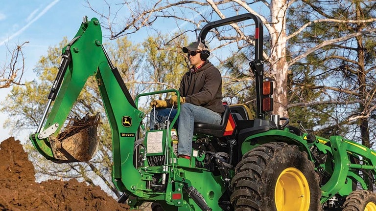 A person with ear and eye protection using a green backhoe to scoop soil.