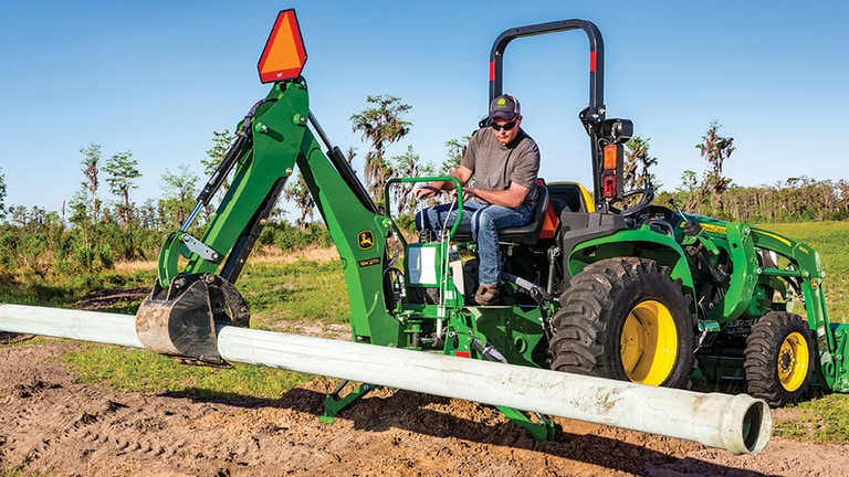 A green backhoe attached to a green compact tractor holds a large pipe above a trench.
