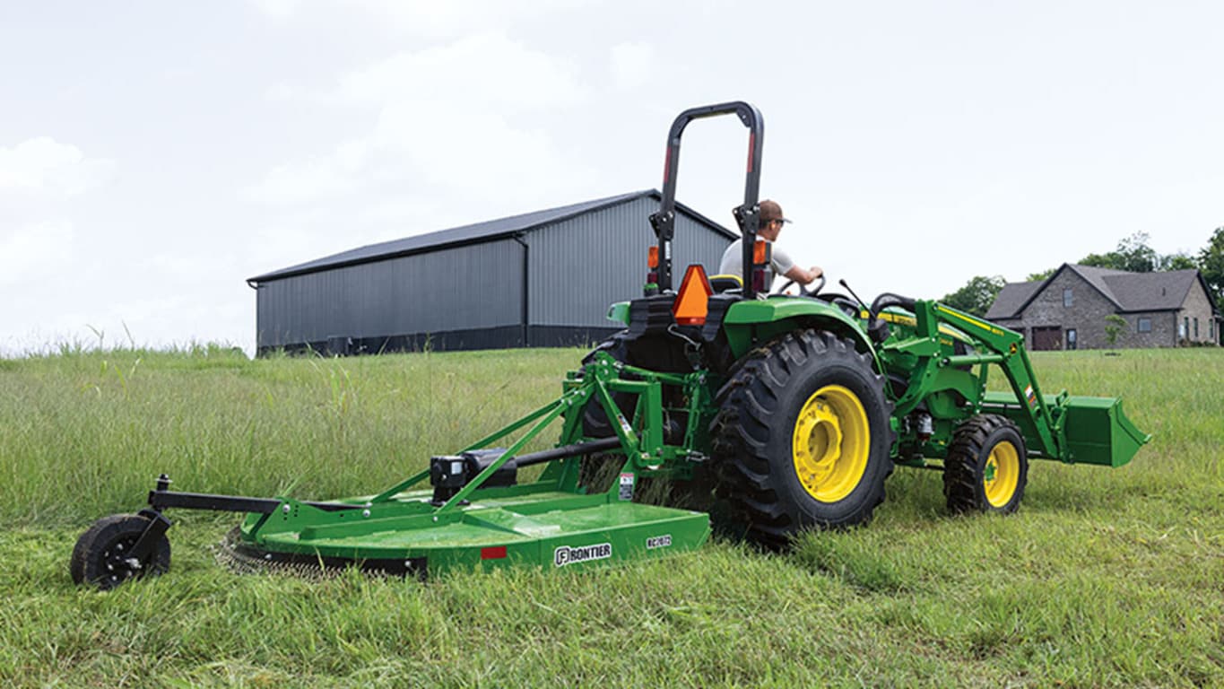 A green compact tractor pulls a rotary cutter through tall grass near a barn.
