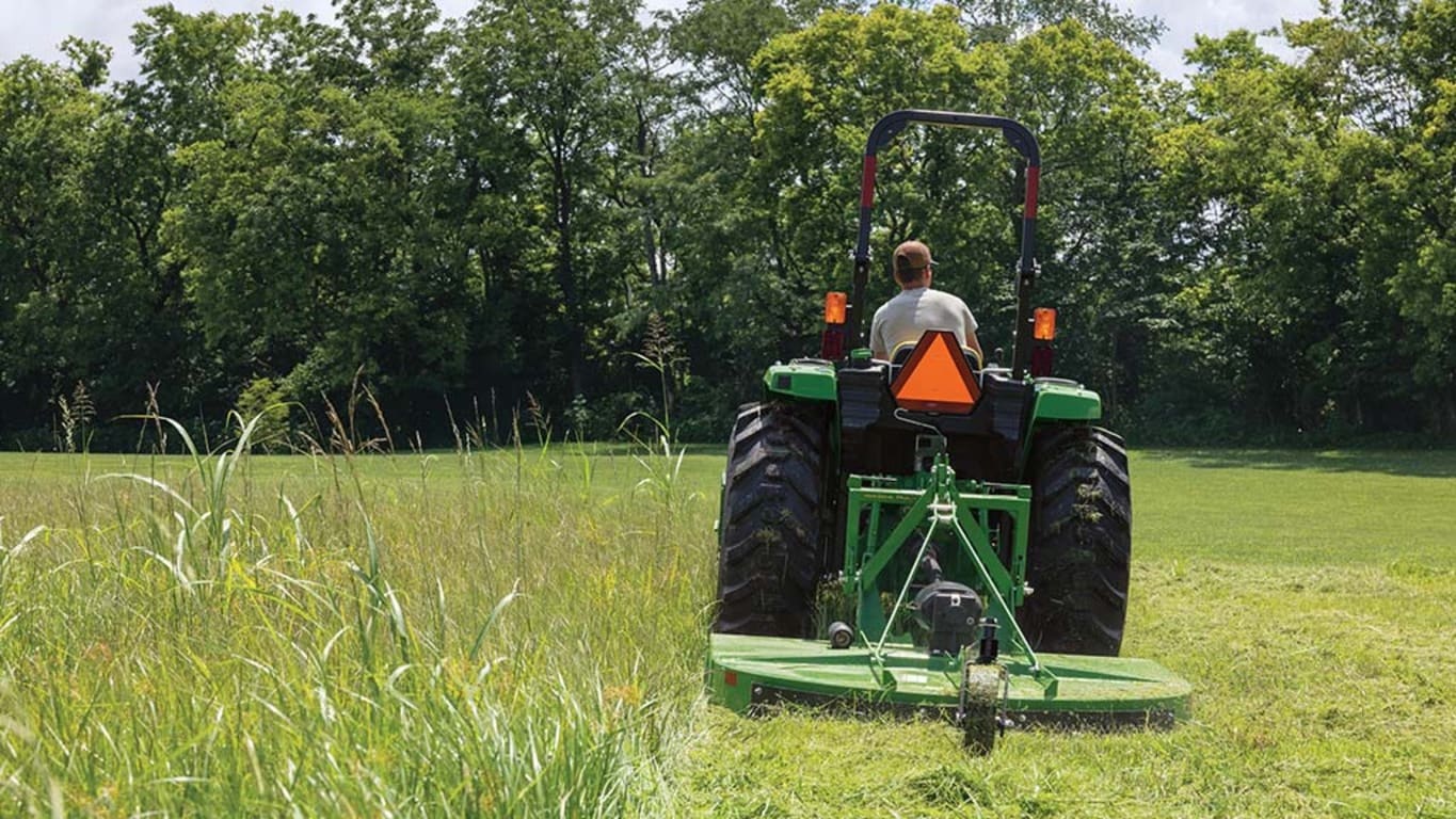 A compact tractor pulls a green rotary cutter through very tall grass in a field bordered by trees.