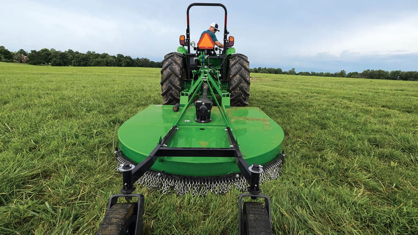 A rotary cutter being pulled by a green compact tractor through a very wide, open field under a cloudy sky.
