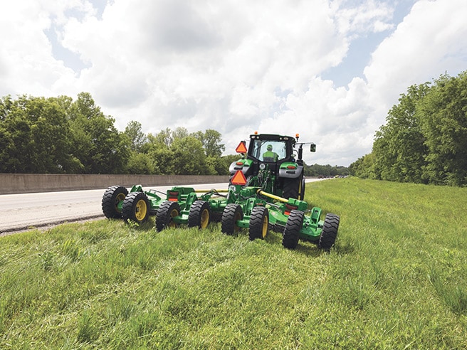 Rear view of green tractor with multi‑deck rotary mower cutting grass along a roadside under cloudy sky