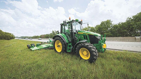 Green tractor towing rotary mower on grassy roadside near highway with concrete barrier and cloudy sky