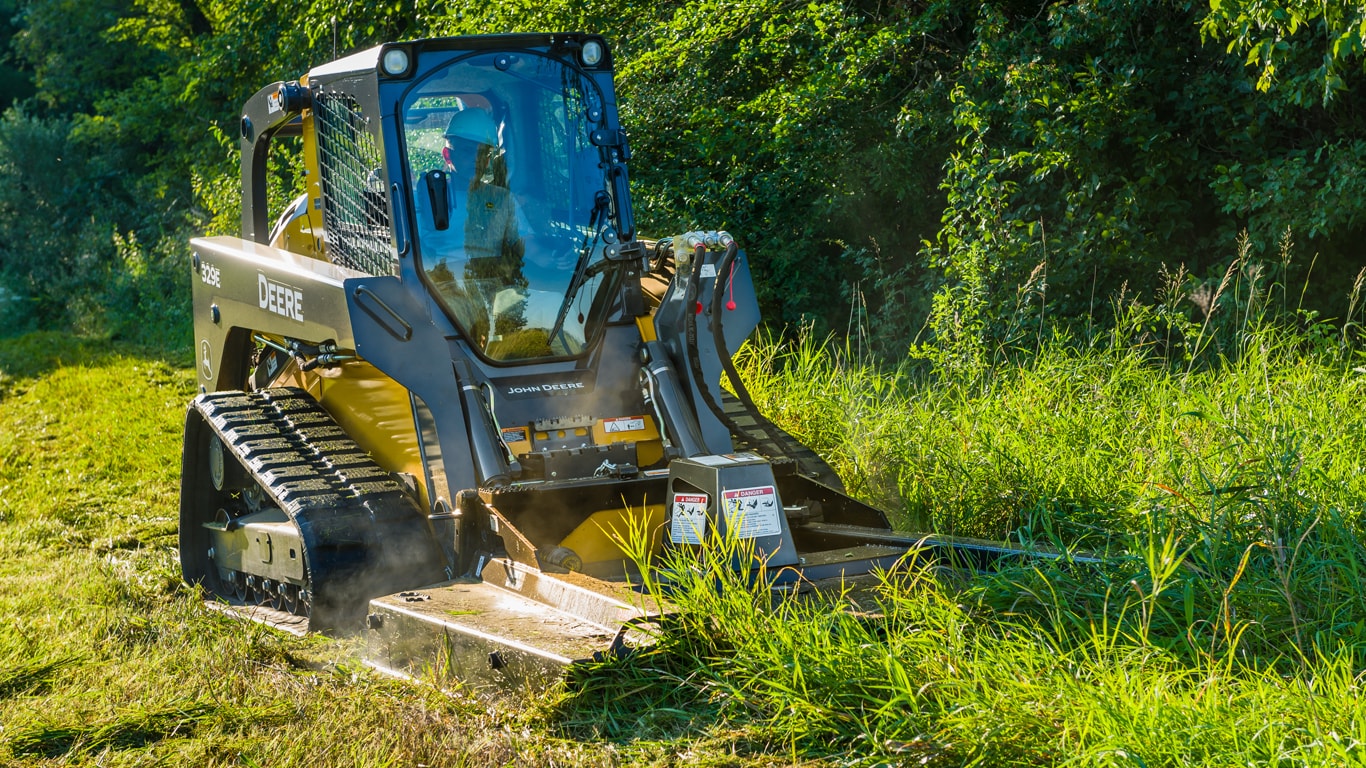 329E Compact Track Loader with RC rotary cutters attachment cutting long grass in a field.