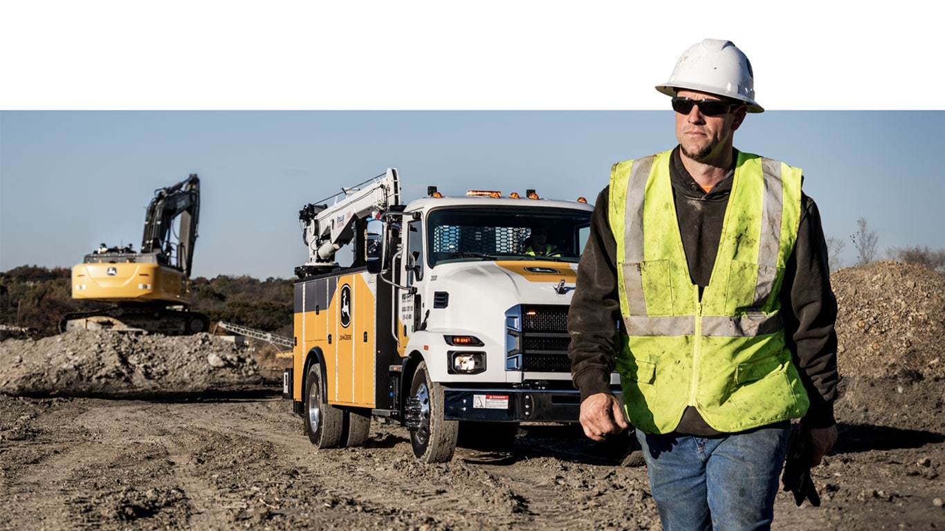 An operator walks in front of a John Deere work truck with an excavator digging in the background.