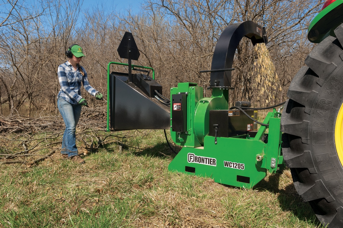 A person in safety gear inserting branches into a green woodchipper surrounded by leafless trees.