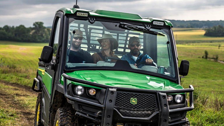 A close-up of a green cabbed Gator carrying three passengers through an open grassy field.