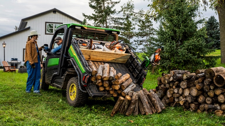 An individual talks to the driver of a Gator through the driver side window while the cargo box tilts up to dump logs.