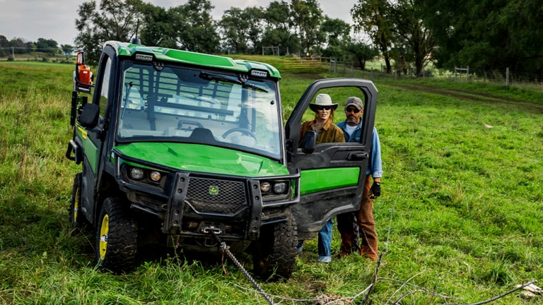 Two individuals stand behind an open driver-side door of a cabbed green Gator while a taut chain is attached to the front.