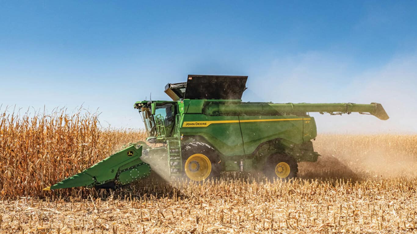 Combine harvester working in a cornfield, with dust rising against a clear blue sky. Harvest in progress.