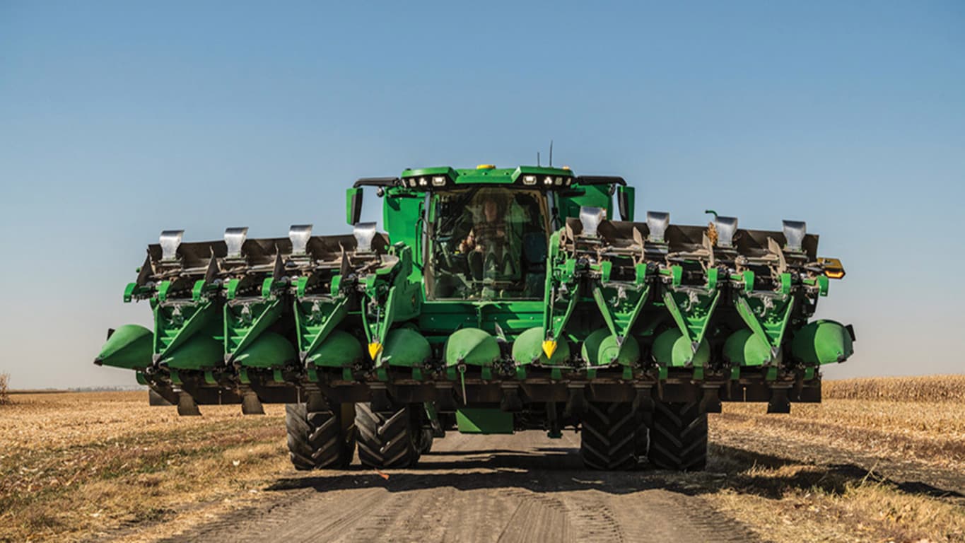 Combine harvester with multiple corn heads moves down a dirt path in a sunny agricultural field.