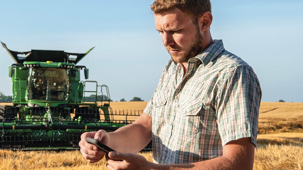Photo of a farmer standing in front of a John Deere S7 Combine that's parked in a wheat field. The farmer is interacting with John Deere Connected Support™ on his mobile device