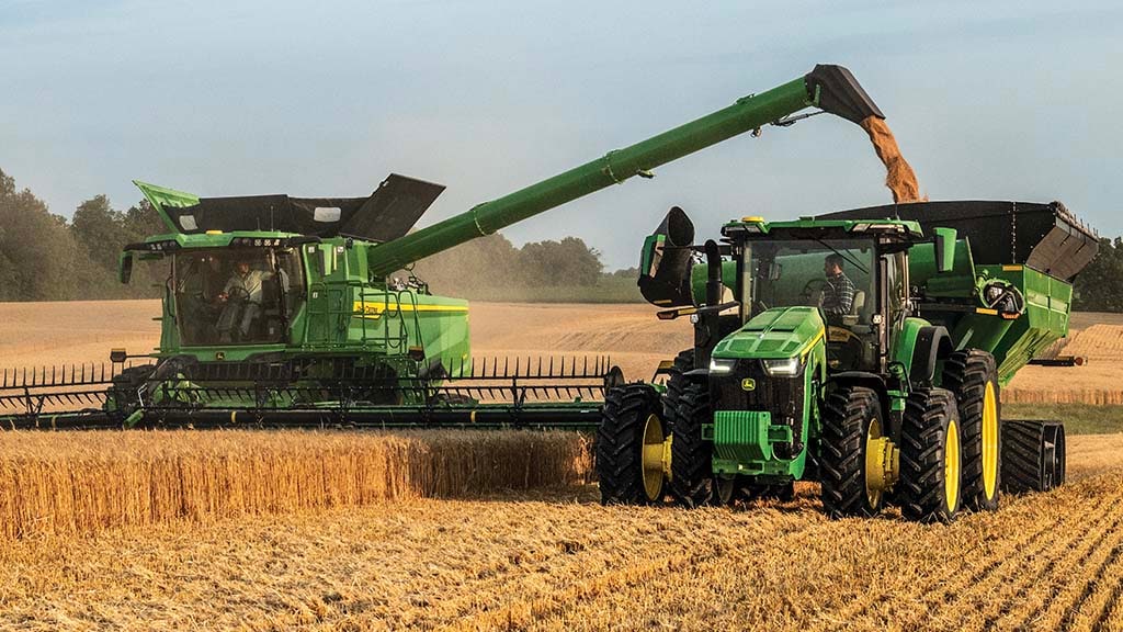 Photo of a John Deere S7 Combine harvesting wheat with a draper head, while unloading grain into a grain cart being pulled by a John Deere tractor