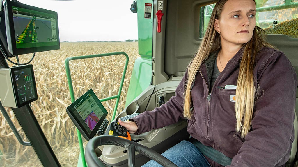 In-cab photo of a young farmer operating a John Deere S7 Combine in corn. You can see before her a G5Plus Display, a New Generation Primary Corner Display and a G5Plus Extended Monitor