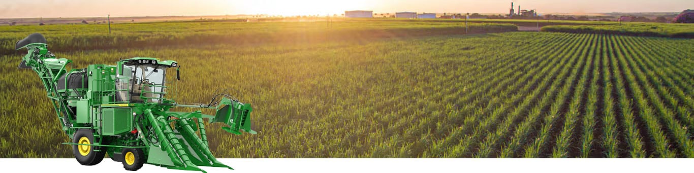 A photo of a partially-harvested sugar cane field with the sun rising on the horizon. A John&nbsp;Deere CH Series Sugar Harvester is inset into the lower left hand corner of the image.