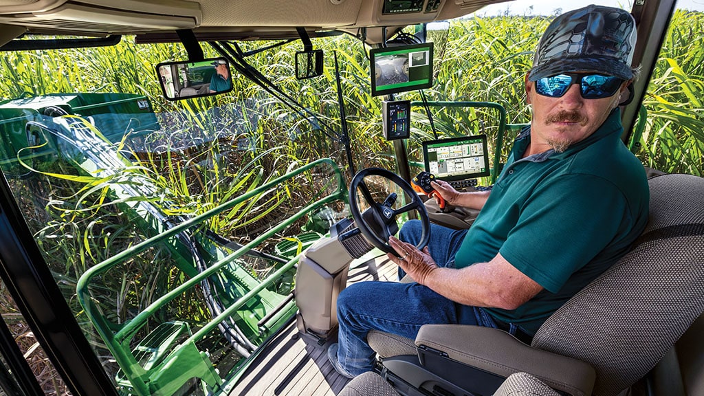 An in-cab, close-up photo showing an operator – in a CH Series Sugar Harvester – turned toward the camera, with standing crops visible through the windows.