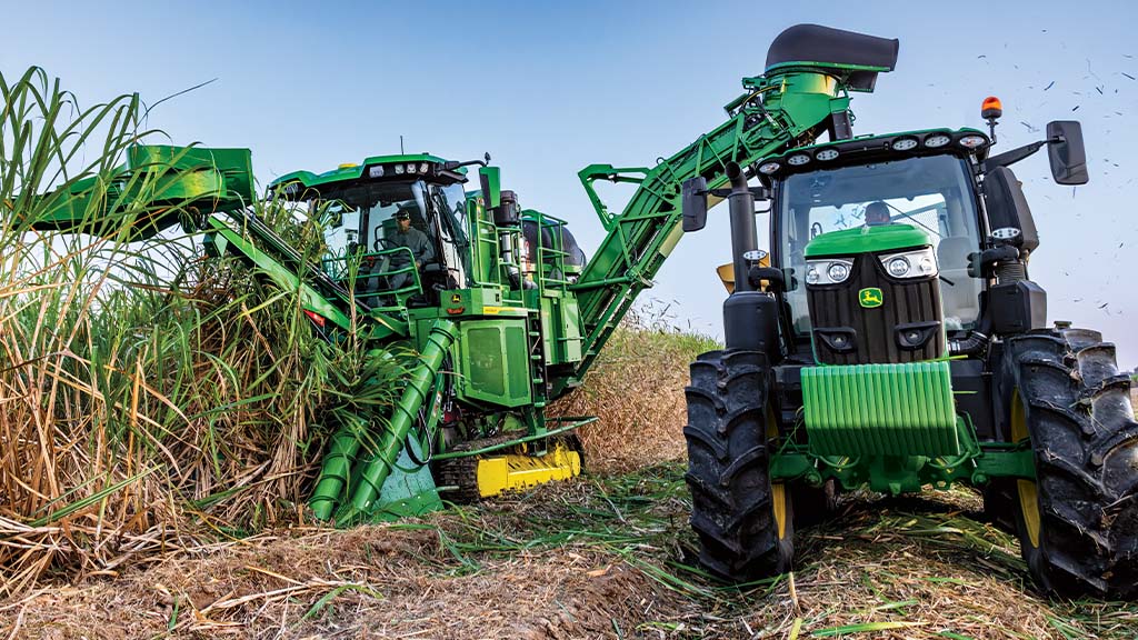 Front-view photo showing a CH Series Sugar Harvester unloading crop into a tractor-pulled grain cart.