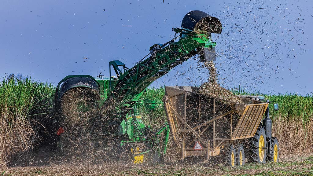 A photo showing CH Series Sugar Harvester unloading sugar cane into a tractor-pulled grain cart, with cane debris floating in the air.