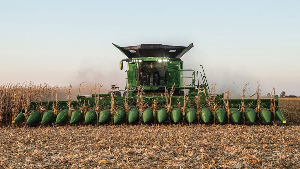 Combine harvester harvesting corn in a field, with corn stalks visible at the front, under a clear sky at dusk.