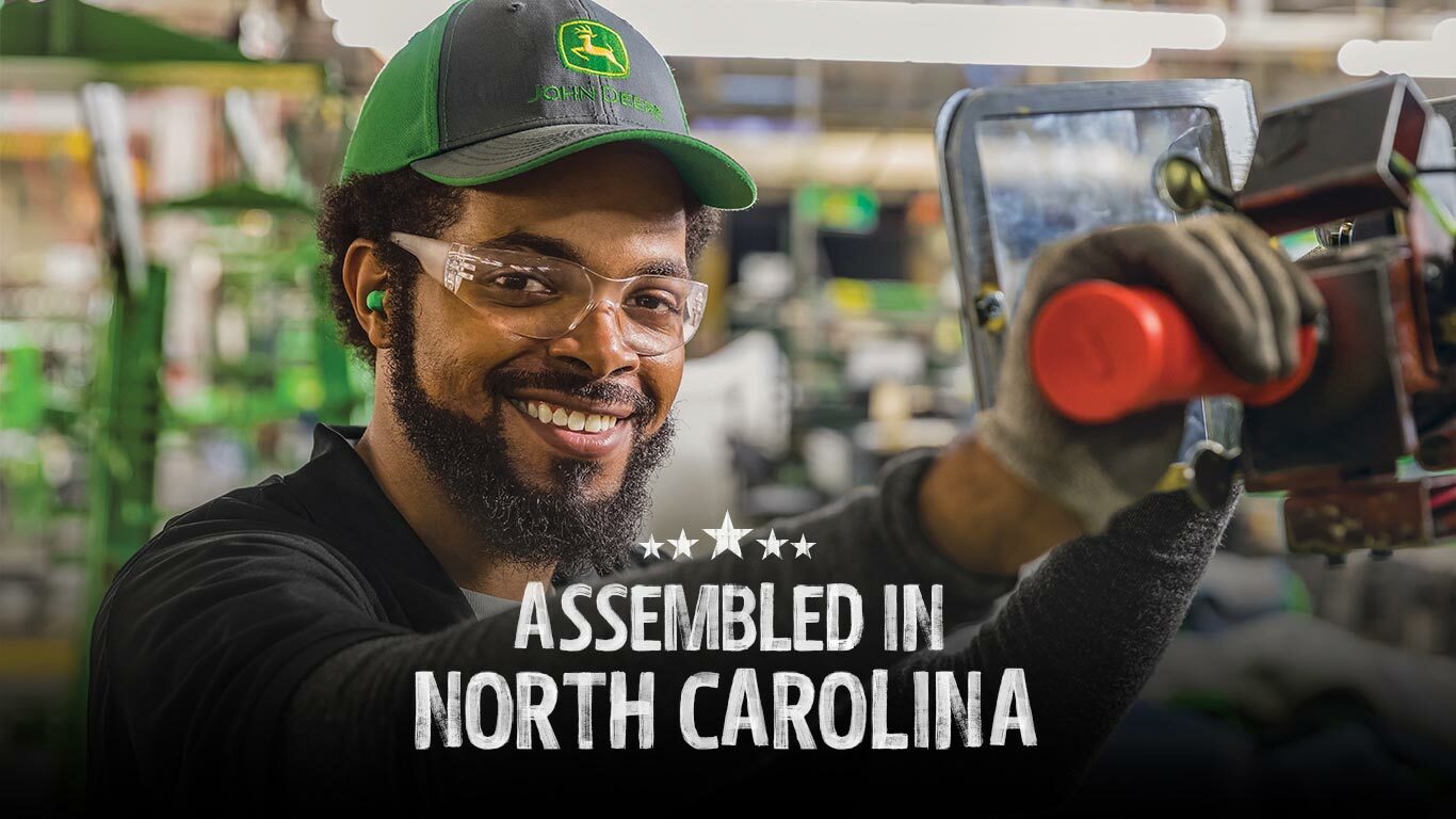 A smiling person safety glasses and a Deere hat assembling a piece of equipment at the factory in North Carolina.