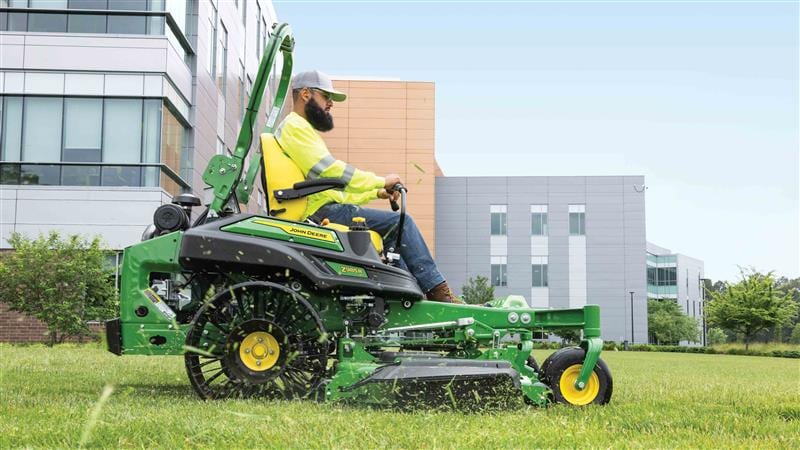 A worker driving a green EFI zero-turn mower with airless tires around a campus.