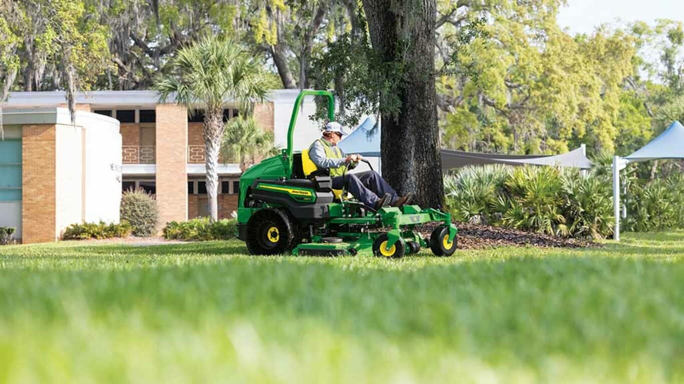 A person in a yellow work vest mowing a college campus lawn with a green zero-turn mower.