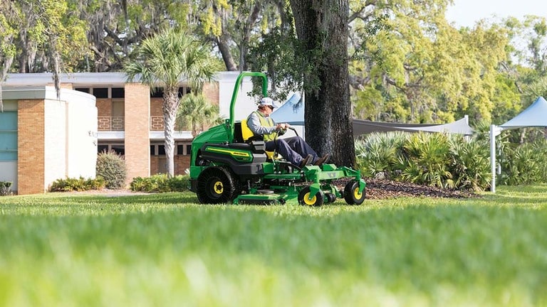 A person in a work vest driving a green zero-turn mower around a mulch patch with a tree in the middle.