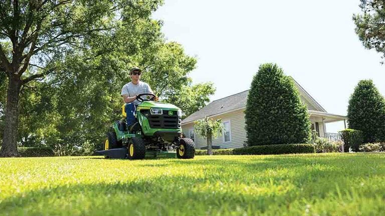 A person driving a green riding mower over a flat open lawn.