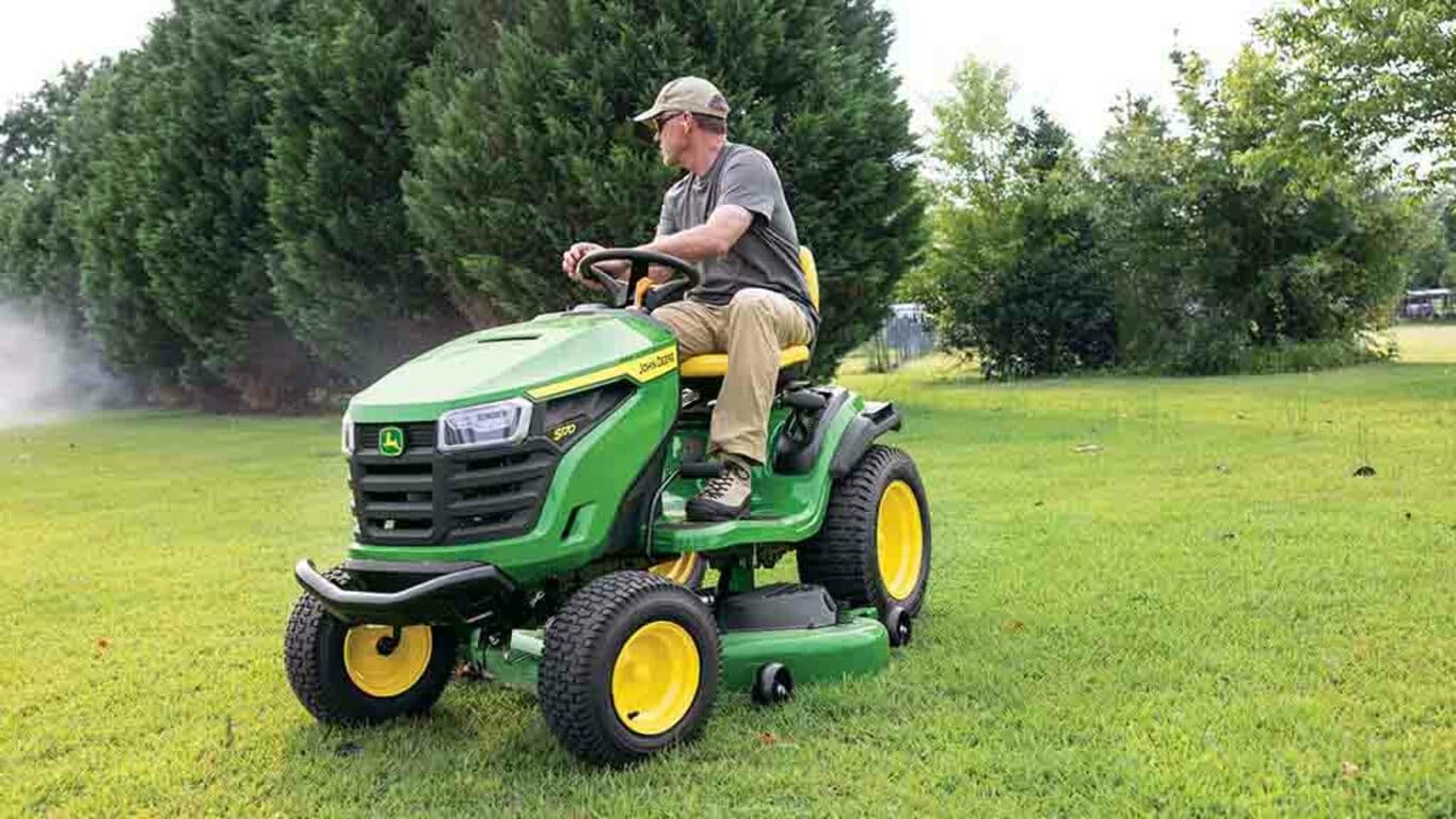 A person turning the steering wheel of a green riding mower in front of large conifers.