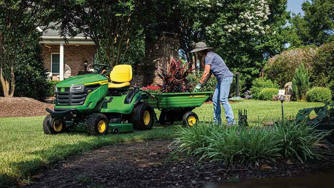 Plants being handled in a green cart attached to the back of a green riding mower.