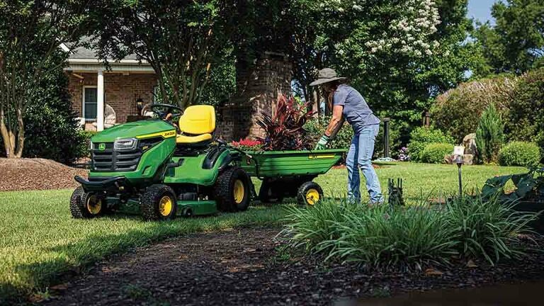 Plants being handled in a green cart attached to the back of a green riding mower.