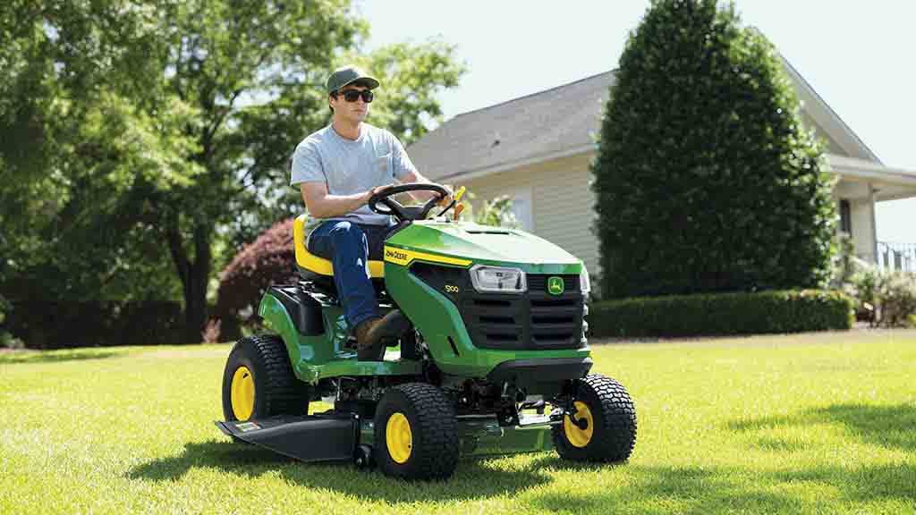 Close-up of a person driving a green riding mower over a flat even lawn.
