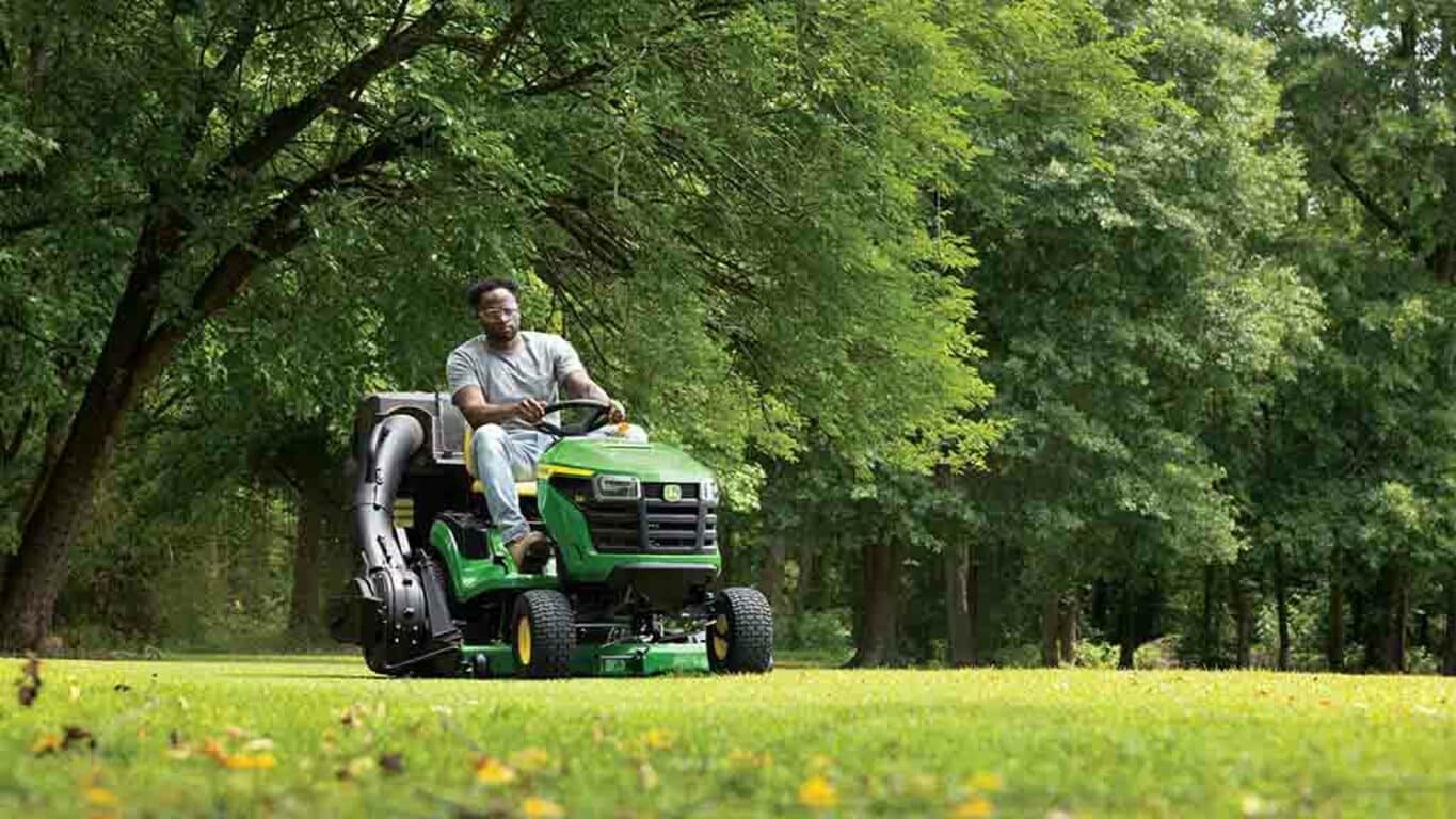 A person driving a green riding mower with a bagging attachment over a well-kept lawn.