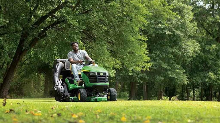 A person driving a green riding mower with a bagging attachment over a well-kept lawn.
