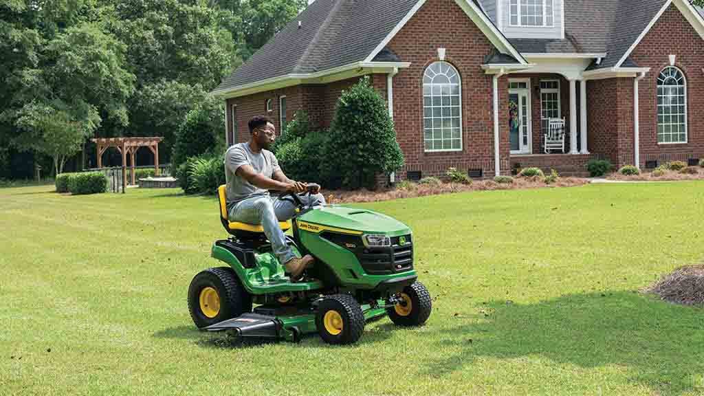 Person driving a green riding mower over a well-kept lawn in front of a home.
