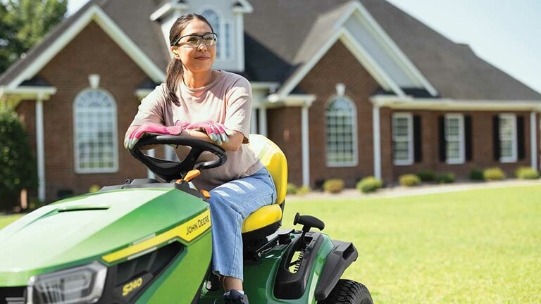 A person smiling while resting on a green riding mower with a on a well-kept lawn.