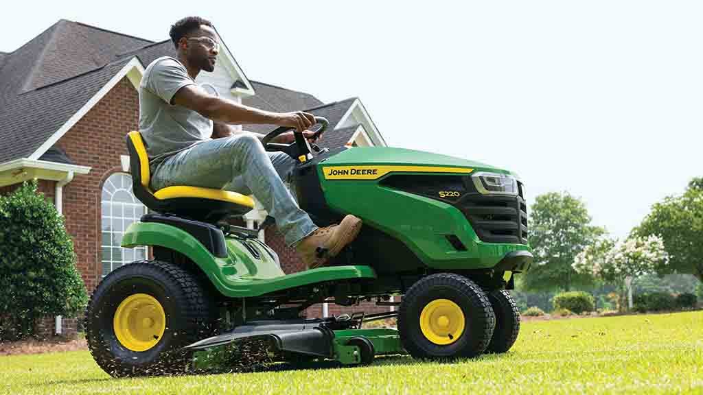 Close-up of a person driving a green riding mower with grass clippings flying out of the deck.