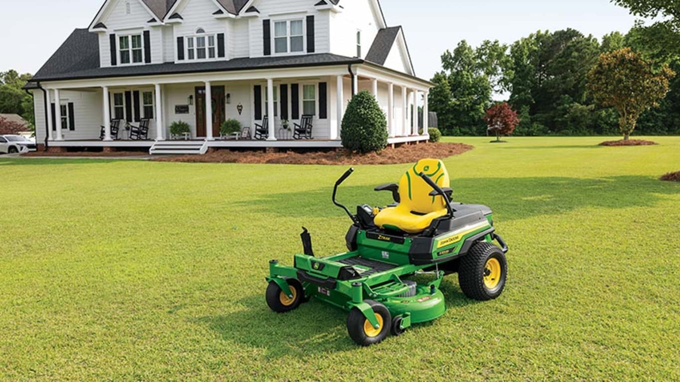 A green zero-turn mower sitting in front of a residence on a well-kept lawn.
