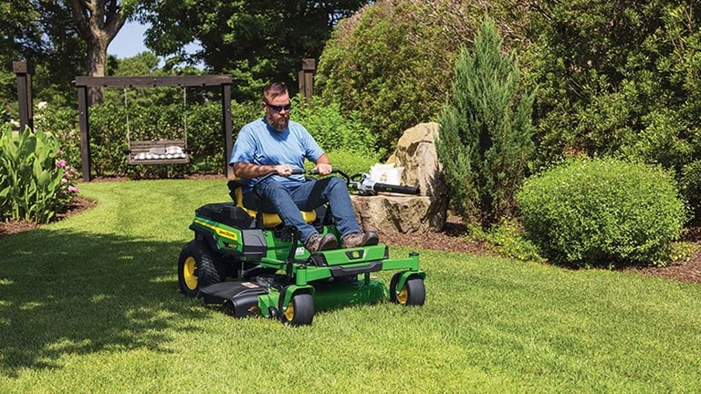A person driving a green zero-turn mower around mulched areas with a swing, bushes, and rocks.