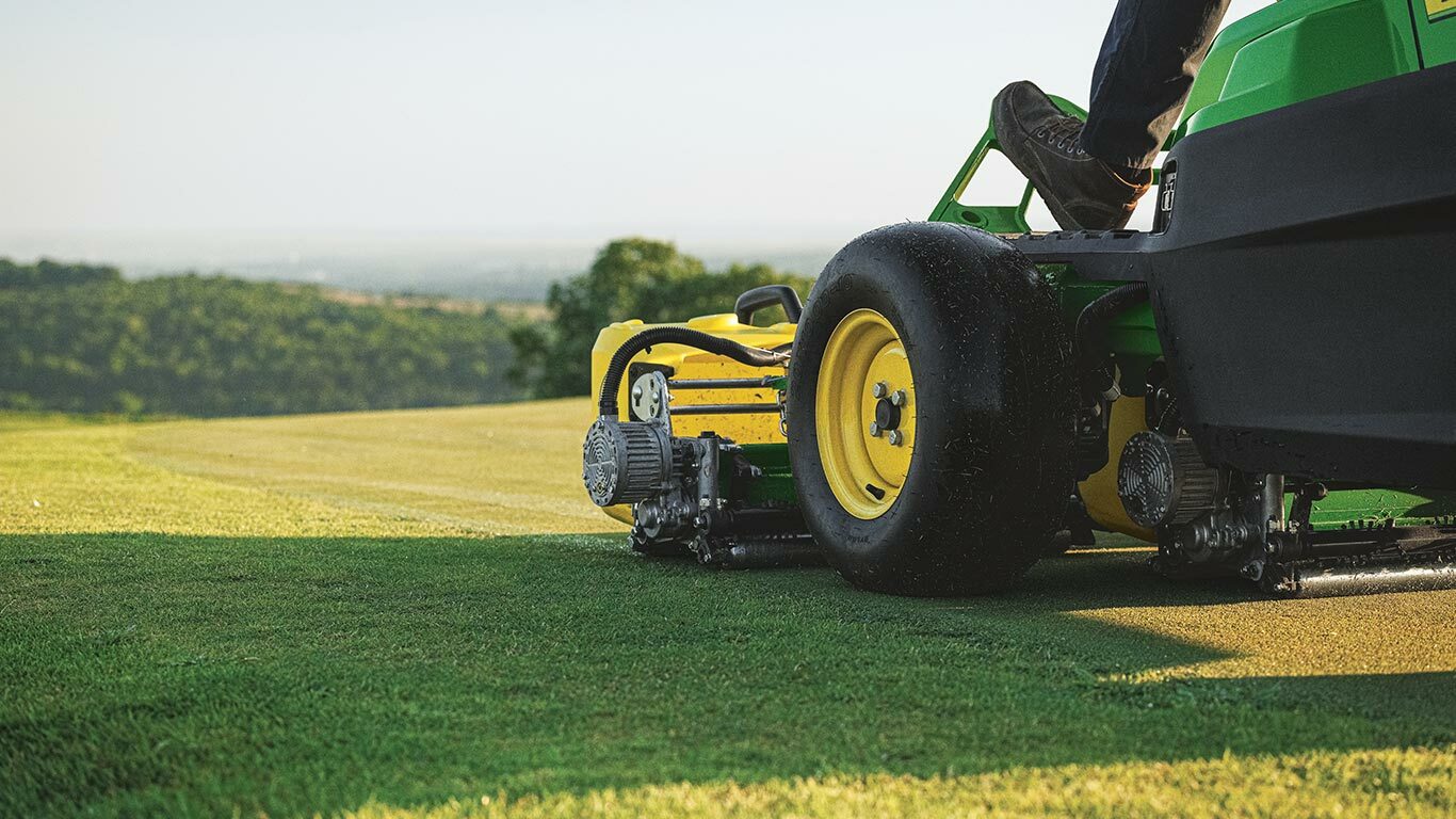 Green and yellow lawn mower cutting grass on golf course with hills and trees in background.