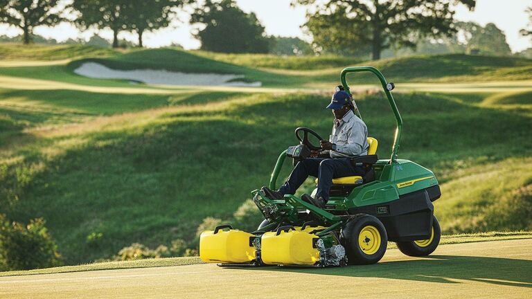 Person driving green John Deere mower on golf course with trees and sand traps in background.