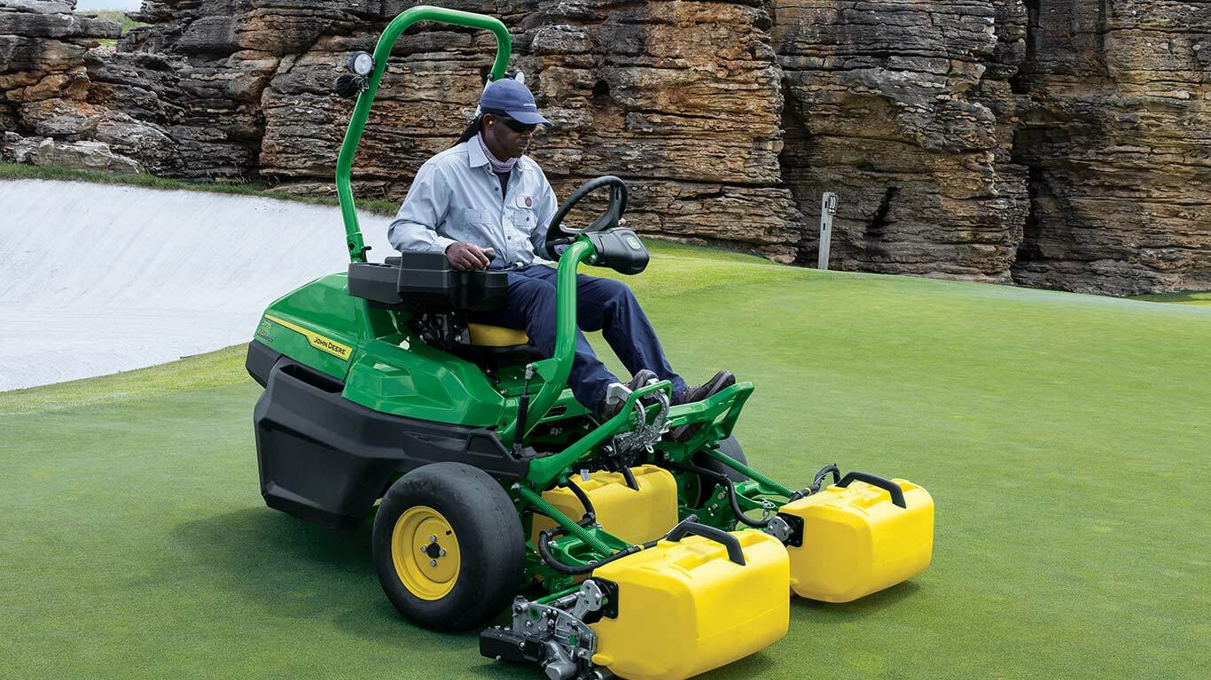Person on green and yellow John Deere mower on golf course near rocky terrain.