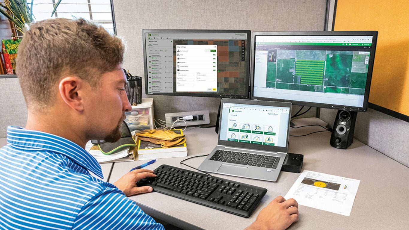 Person at a desk in front of dual computer screens and laptop looking at operations center screens.