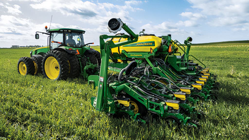 A green John Deere tractor with a planting attachment is shown in a field, preparing soil under a partly cloudy sky.