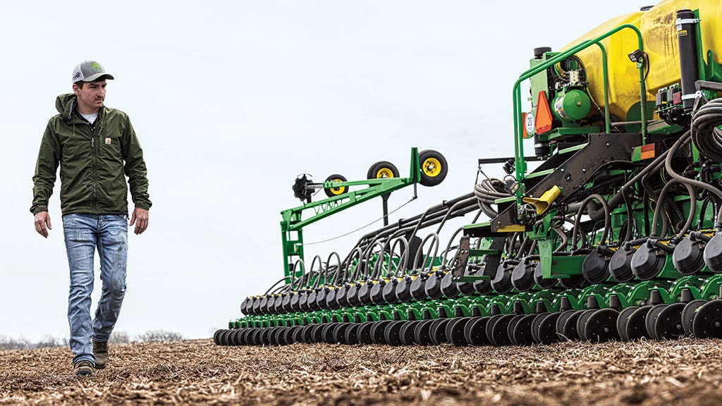A person walks alongside agricultural machinery on a field, with rows of planting equipment and a cloudy sky in the background.