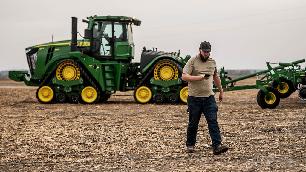 Person using a smartphone in a field with a tracked tractor and tillage equipment in the background.