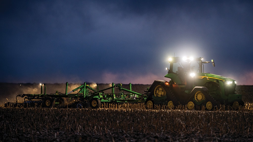 Tractor pulling tillage equipment at night with headlights illuminating a harvested field.