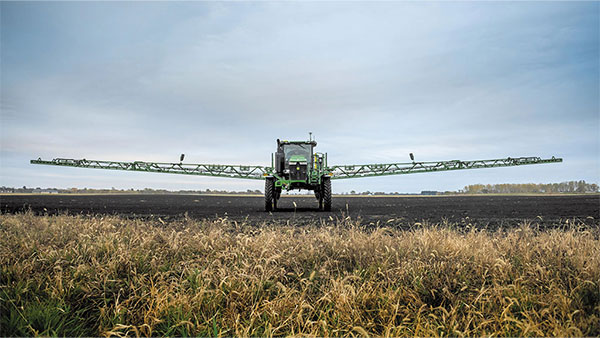 Rear view of a John Deere self-propelled sprayer with fully extended boom arms crossing a dark field under an overcast sky.