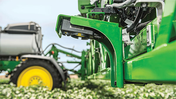 Close-up of a John Deere sprayer boom section with mounted sensor nozzle components, with a tractor blurred in the background over a low crop field.